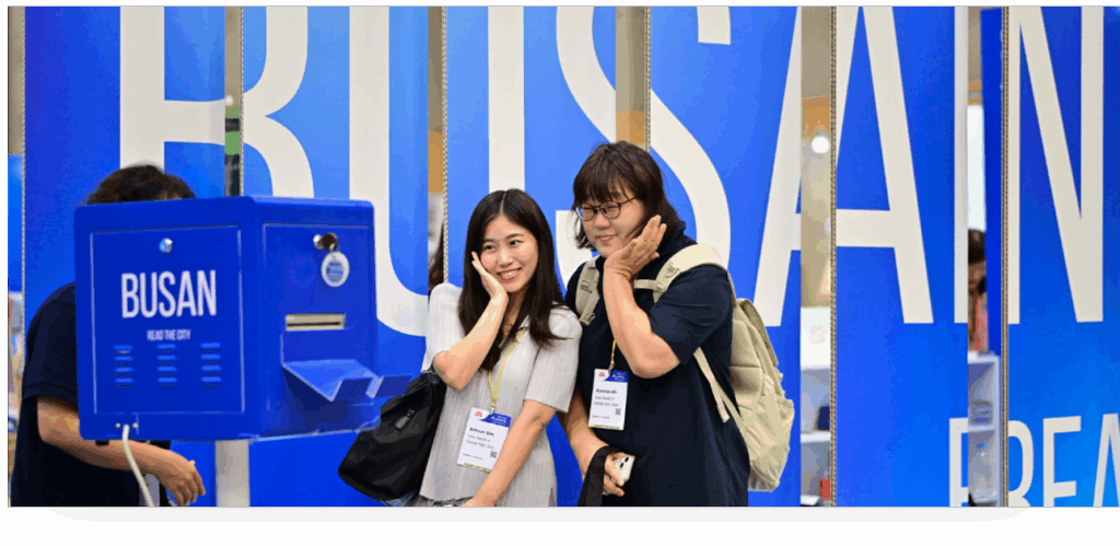 2 asiatisch gelesene Frauen posieren vor einer Kamera, auf der BUSAN steht